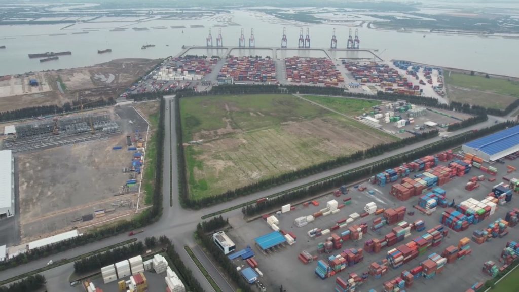 Bãi container và khu đất trống dọc trục đường trong khu công nghiệp ven sông nhìn từ trên cao, phía xa là hàng cẩu bến cảng