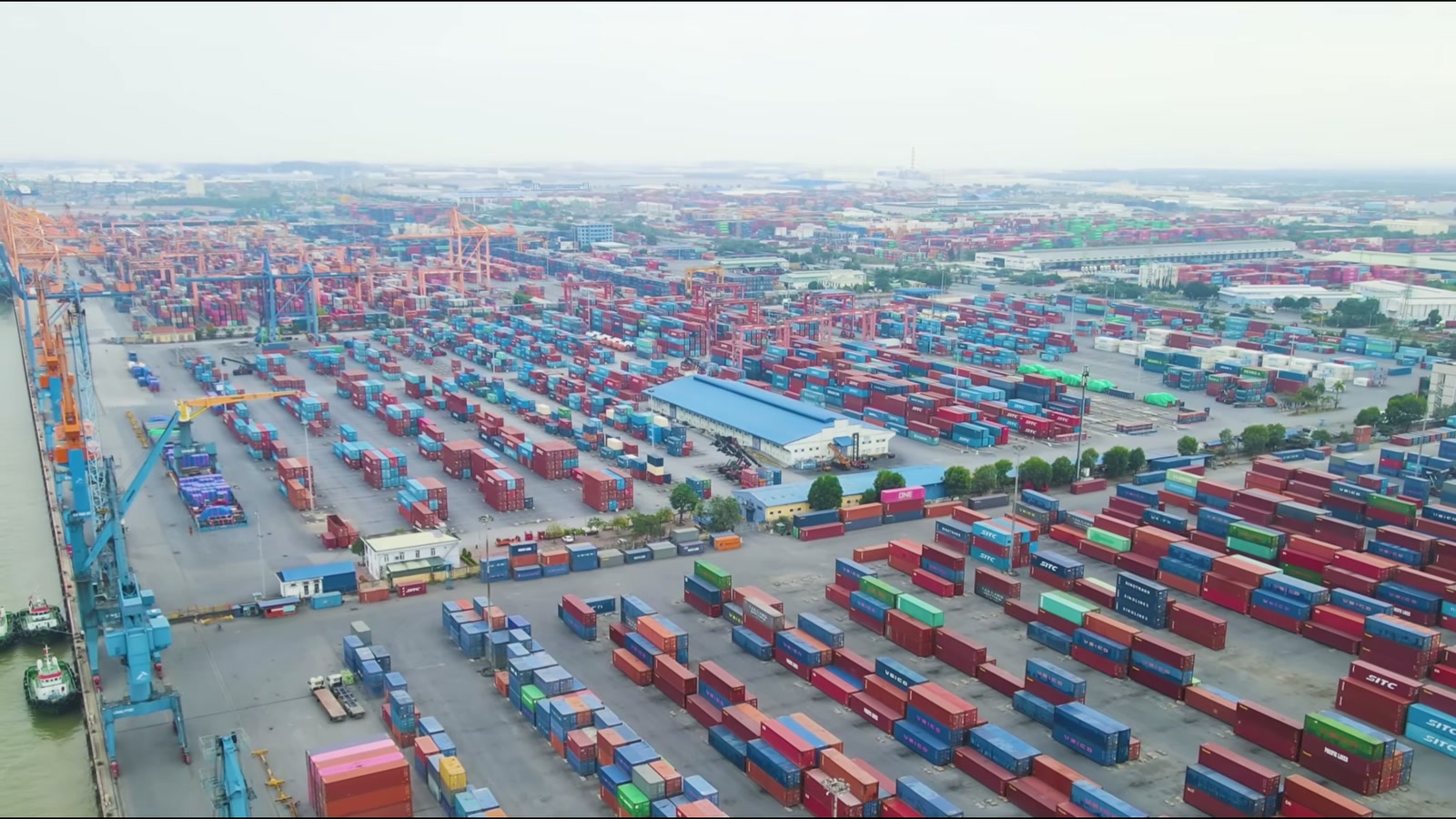 Bãi container xếp thành nhiều dãy dài nhìn từ trên cao, cần cẩu bờ màu xanh đứng sát mép nước, xe đầu kéo chạy giữa các lối đi
