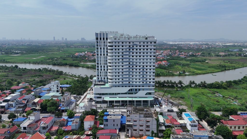 Bệnh viện Đa khoa Quốc tế Thủy Nguyên nhìn từ trên cao, toà tháp đứng giữa khu dân cư mái đỏ và dòng sông uốn quanh phía sau