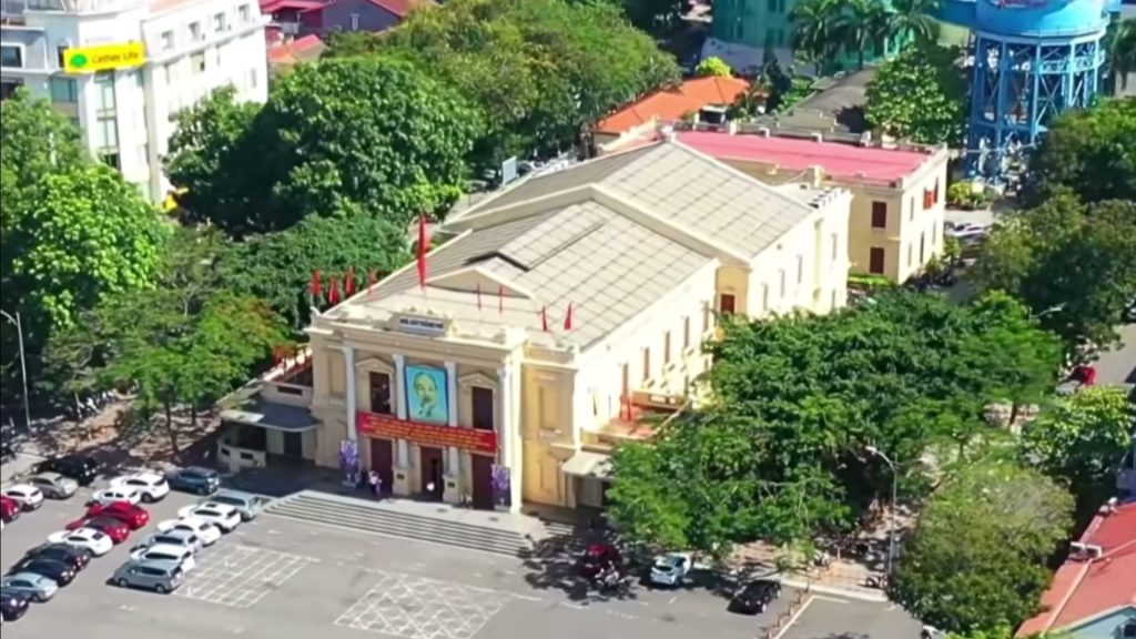 Nhà hát lớn Hải Phòng nhìn từ trên cao, toà nhà kiểu Pháp màu vàng bốn mặt tiền giữa hàng cây xanh và bãi đỗ xe