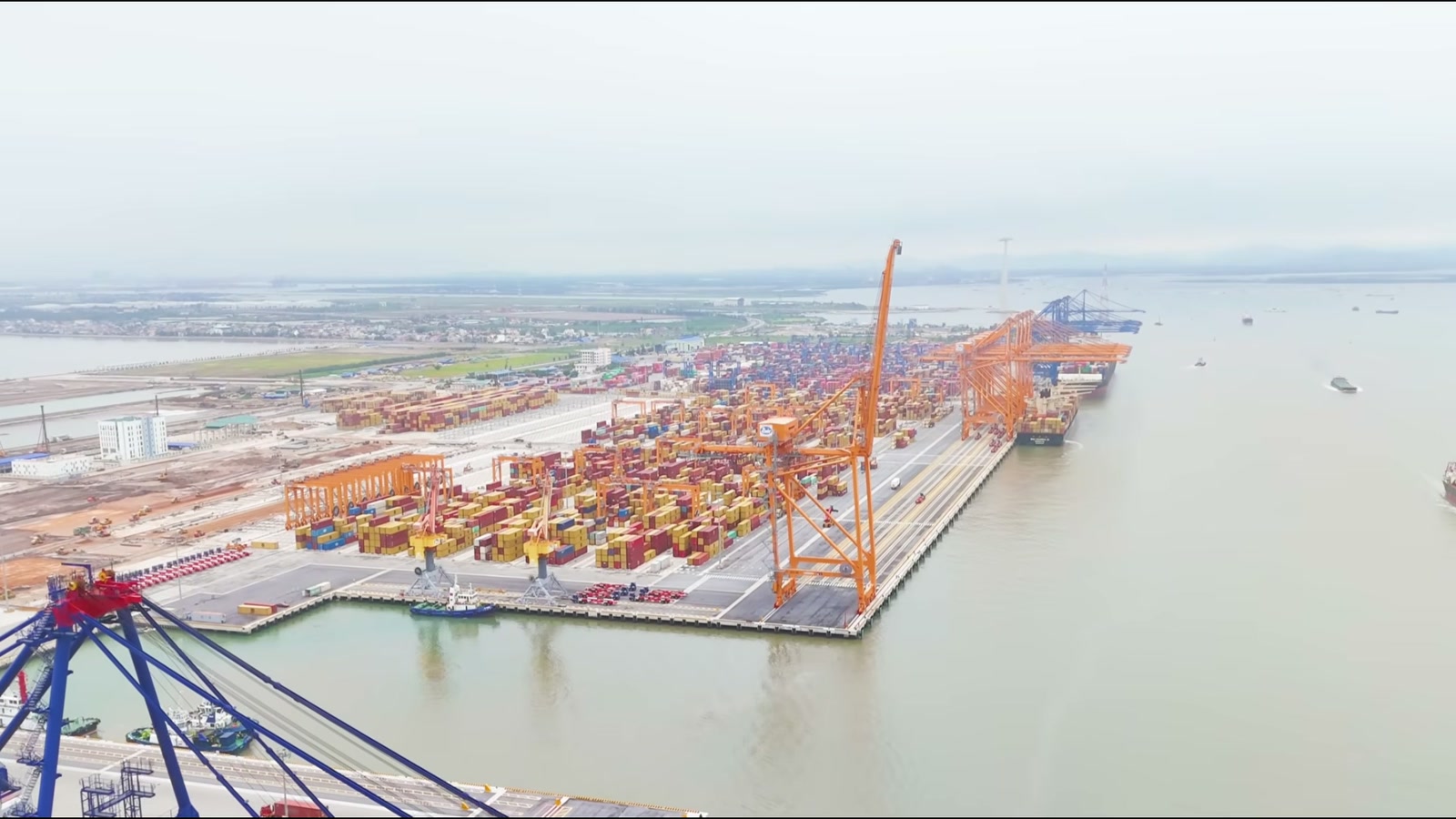 Cảng container ven sông nhìn từ trên cao, hàng cần cẩu màu cam đứng dọc cầu cảng, bãi container nhiều màu phía sau và tàu hàng neo ngoài luồng
