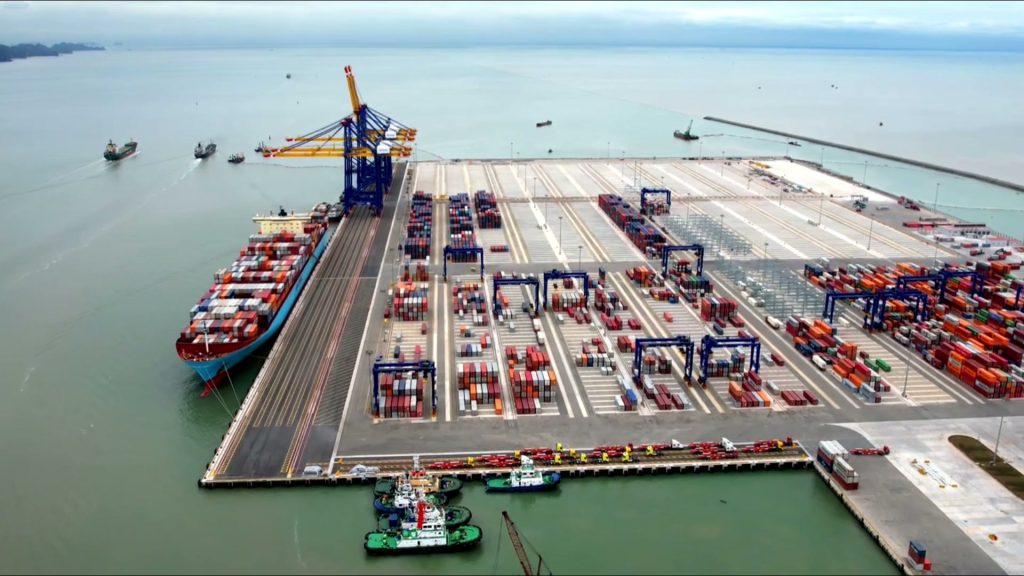 Bến cảng container nước sâu nhìn từ trên cao, tàu container lớn cập cầu cảng, hàng cẩu bờ màu xanh vàng và bãi container xếp nhiều màu, tàu kéo neo phía trước