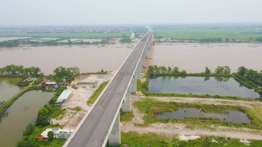 Cầu cạn bê tông băng qua một con sông rộng và vùng ruộng ao nhìn từ trên cao, đường dẫn vươn thẳng về phía đồng bằng xa