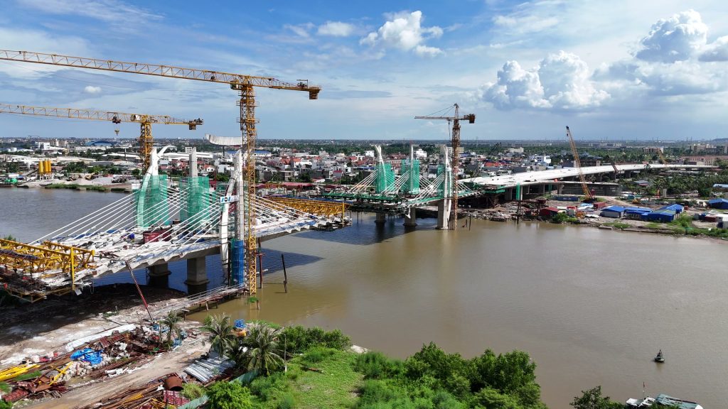 Công trường cây cầu dây văng đang thi công bắc qua sông ở Hải Phòng nhìn từ trên cao, hai cặp trụ tháp bọc lưới xanh, cần cẩu tháp và các nhịp cầu vươn ra từ hai bờ dưới trời xanh mây trắng