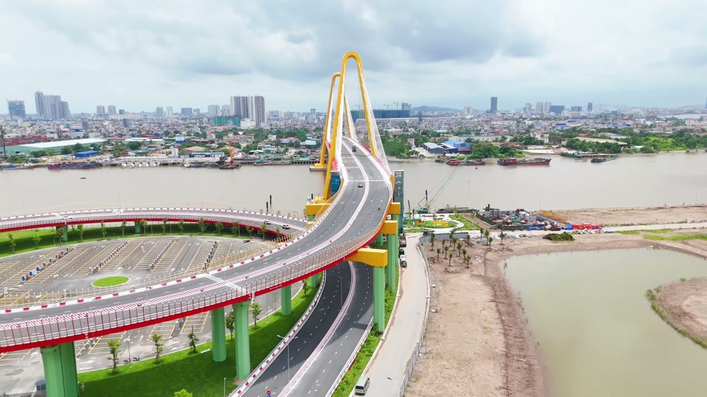 Cầu Hoàng Gia bắc qua sông Cấm nhìn từ trên cao, đường dẫn uốn vòng màu đỏ trên hàng trụ xanh, hai trụ tháp vàng hình cánh cổng, phía xa là nội thành Hải Phòng