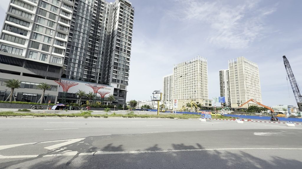 Cụm chung cư cao tầng đã hoàn thiện đứng cạnh một công trường quây rào bên trục đường lớn, phía xa có cần cẩu tháp