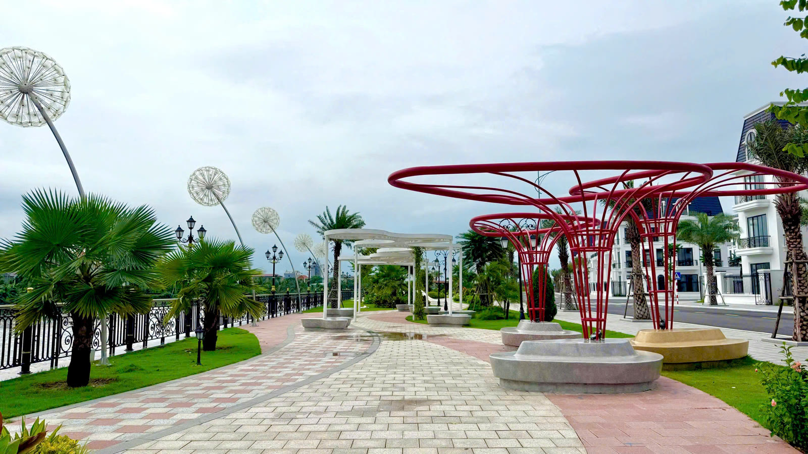 Lối đi lát gạch ven sông trong công viên Golden Park, giàn hoa sắt màu đỏ, đèn trang trí hình bồ công anh và hàng cọ, phía sau là dãy nhà phố