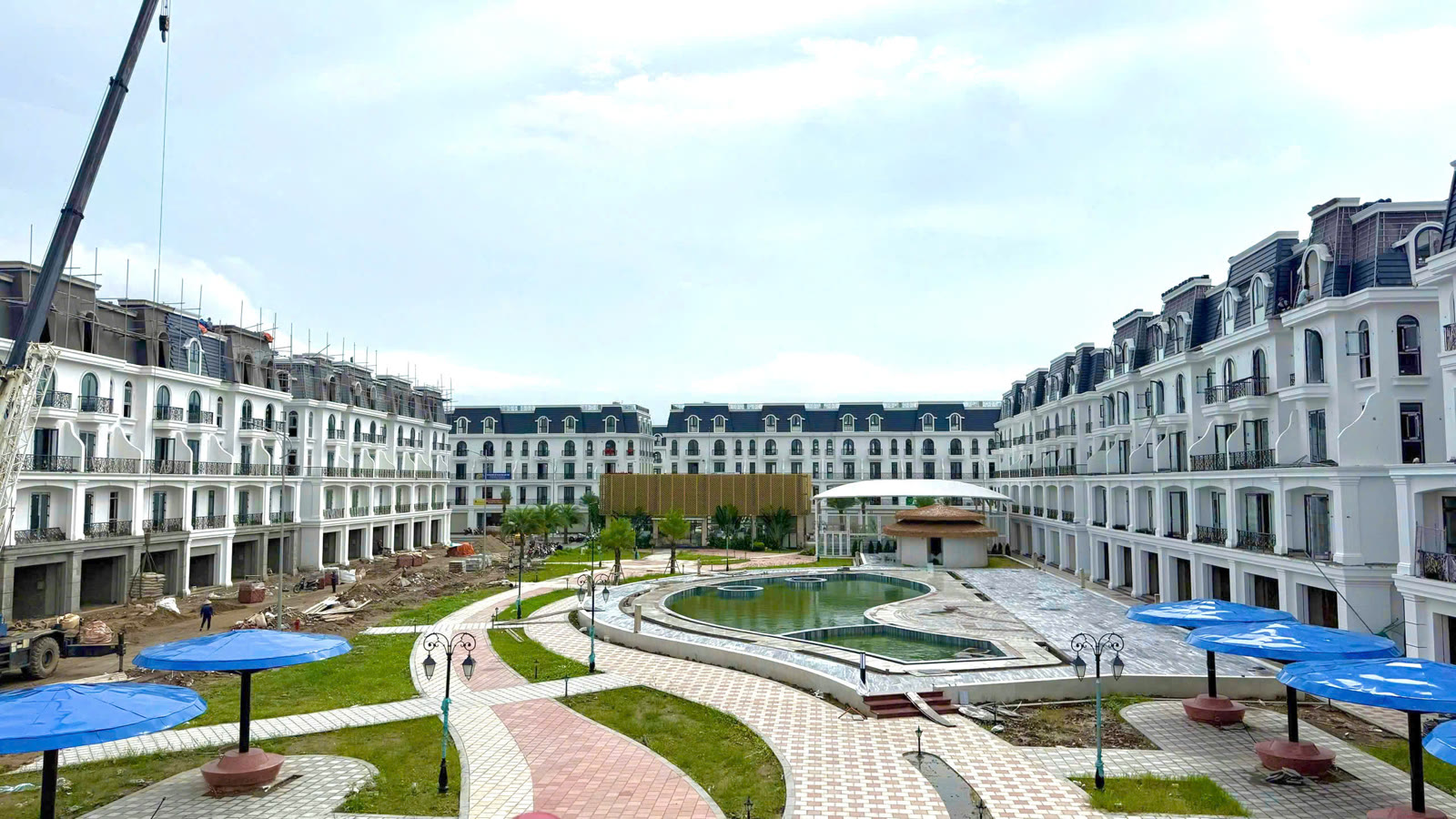 Công viên Joy Garden Park với bể bơi uốn cong ở giữa, các ô dù màu xanh dọc lối đi, bao quanh là dãy nhà phố kiến trúc Pháp màu trắng