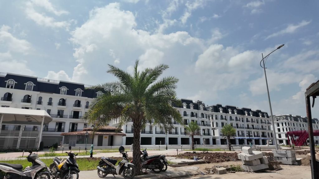 Dãy liền kề phân khu Central Park nhìn từ bãi đỗ xe, nhà đã lên mái, phía trước là cây cọ và vài xe máy dựng ven đường