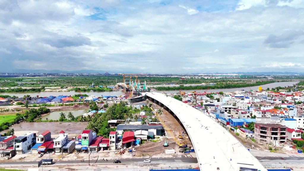 Đường dẫn cầu Hải Thành bằng bê tông uốn cong qua khu dân cư mái đỏ, phía xa là trụ tháp cầu bên sông Lạch Tray