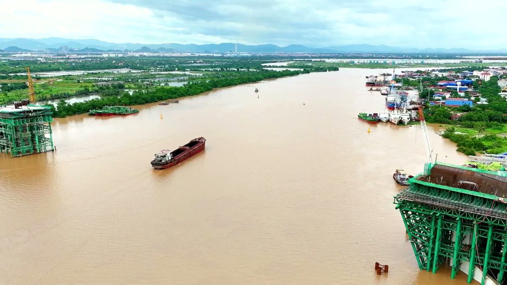 Hai bệ trụ tháp cầu Nguyễn Trãi đứng hai bên lòng sông Cấm, một sà lan chạy giữa sông, phía xa là khu dân cư ven sông và dãy núi