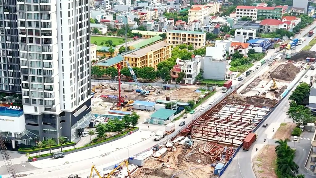 Hệ khung chống thép màu đỏ dựng trong lòng hố đào hầm chui nhìn từ trên cao, xe container và xe tải chạy trên làn đường tạm sát mép công trường, bên trái là một toà chung cư