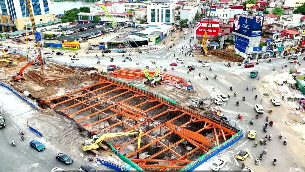 Hố đào hầm chui với hệ văng chống thép màu cam nằm giữa một nút giao đông xe trong khu trung tâm, máy xúc và cần cẩu làm việc bên trong rào chắn