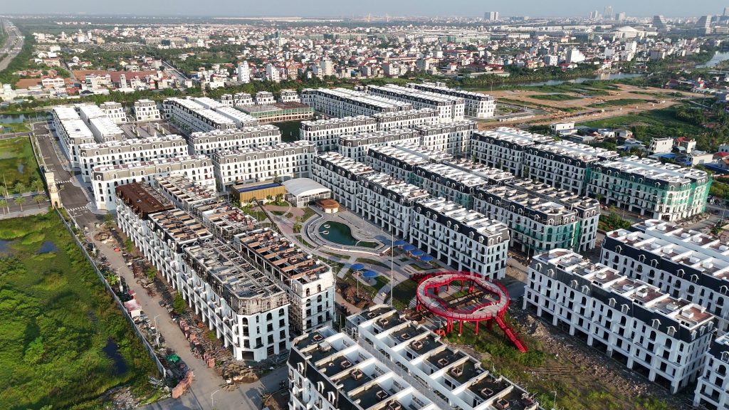 Khu nhà phố mái đỏ xây thành từng dãy dài quanh một công viên có vòng xoay đỏ, nhìn từ trên cao, phía sau là cánh đồng và khu dân cư cũ