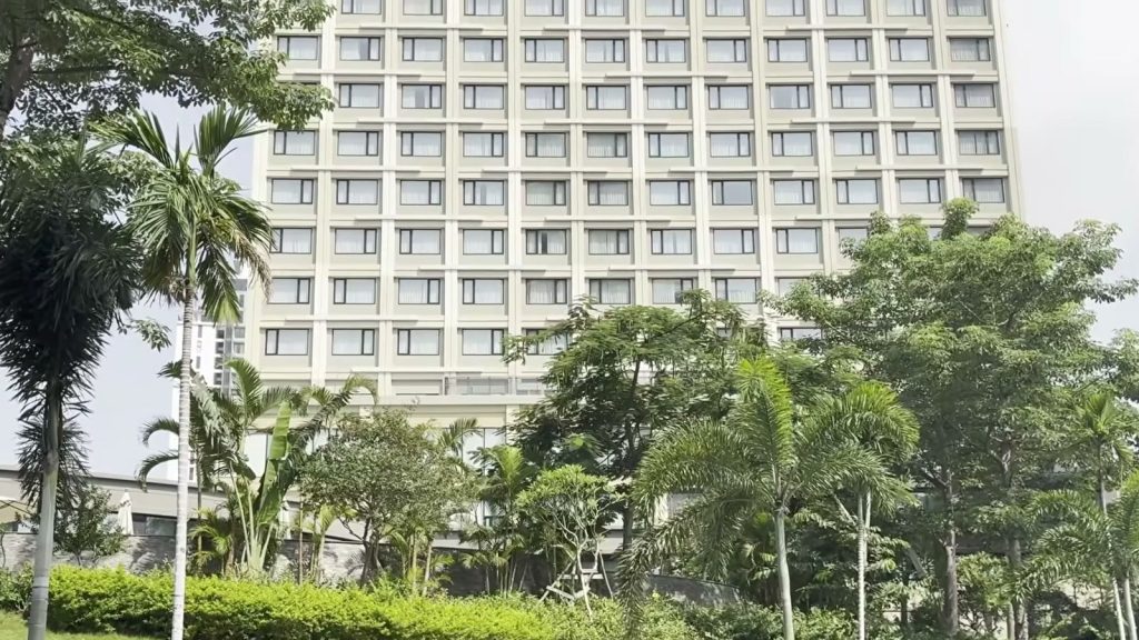 Mặt tiền khách sạn Nikko Hải Phòng màu kem với các ô cửa sổ vuông đều nhau, phía trước là vườn cây xanh, hàng cau và bãi cỏ dốc