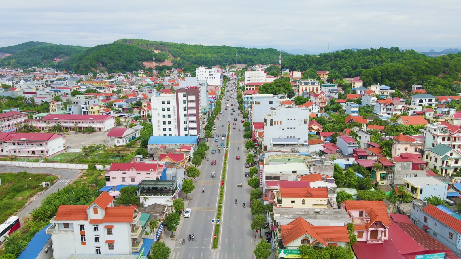 Khu phố hai bên một trục đường thẳng có dải cây xanh ở giữa, nhà mái đỏ xen nhà cao tầng, phía xa là dãy đồi phủ rừng