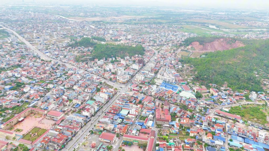 Khu phố Núi Đèo nhìn từ trên cao, nhà mái đỏ và mái tôn xanh dày đặc quanh trục đường chính, bên phải là đồi cây và một vạt núi đất đỏ