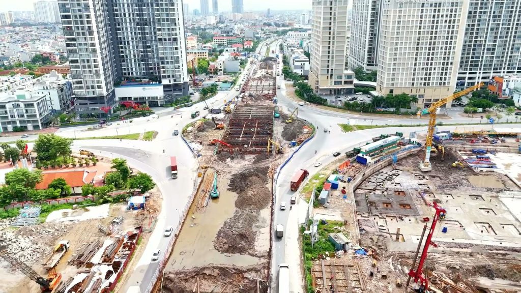 Công trường hầm chui nhìn từ trên cao với hố đào chạy dọc trục đường, cần cẩu và máy khoan cọc, xe cộ lưu thông quanh một nút giao vòng xuyến ở Hải Phòng, phía sau là các toà chung cư