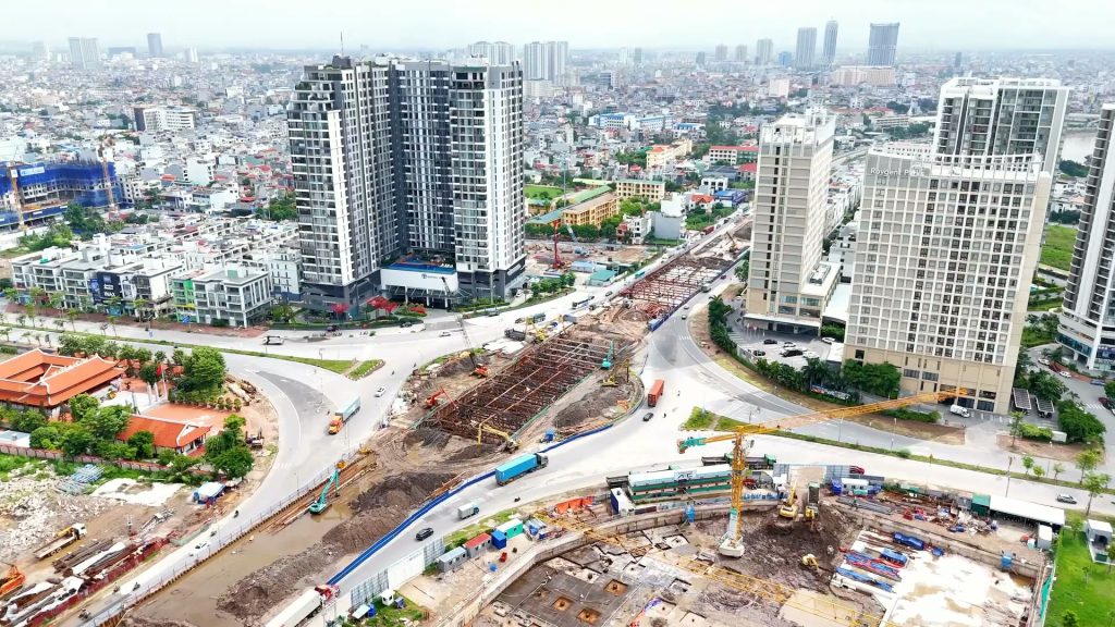 Công trường hầm chui chạy dọc một trục đường lớn cắt qua nút giao vòng xuyến ở Hải Phòng nhìn từ trên cao, xe container chạy trên làn tạm, phía sau là các toà chung cư cao tầng