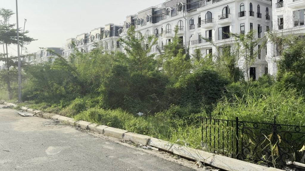 Dãy liền kề phân khu Central Park còn đang thi công nhìn từ mép đường, phía trước là lô đất trống cỏ mọc cao và hàng rào sắt