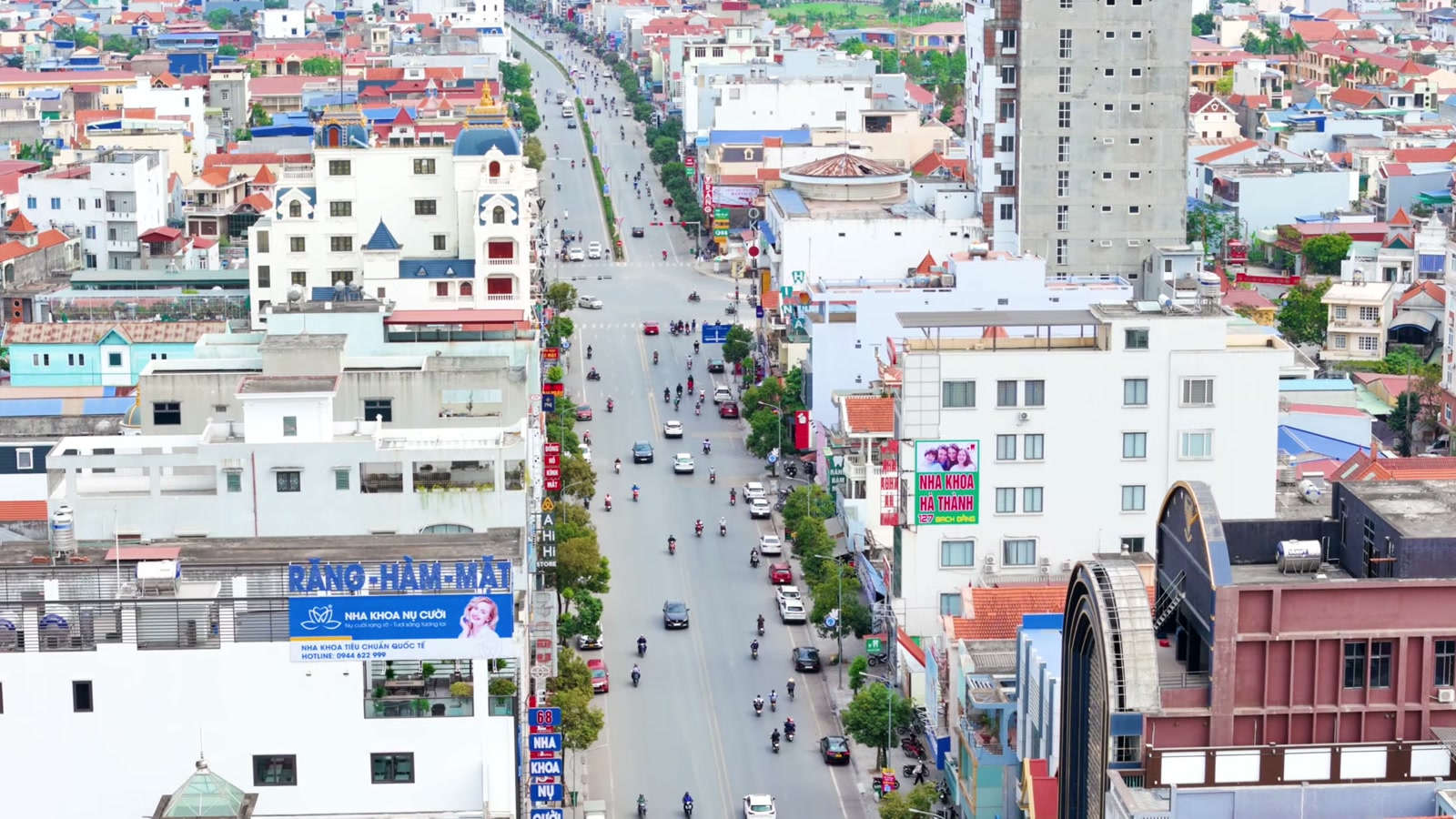 Phố buôn bán đông xe máy nhìn từ trên cao, hai bên là nhà phố nhiều màu và vài toà nhà cao tầng đang xây