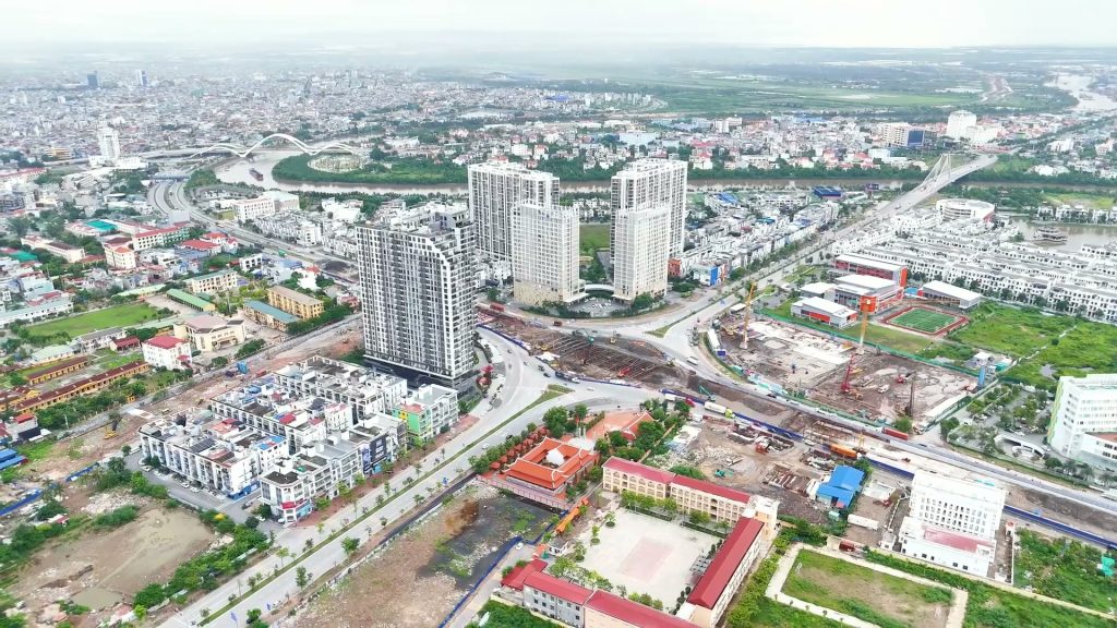 Toàn cảnh nút giao vòng xuyến đang thi công hầm chui ở Hải Phòng nhìn từ trên cao, xung quanh là các toà chung cư và trường học, phía xa là dòng sông với một cây cầu vòm và một cây cầu dây văng