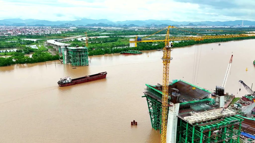 Bệ trụ tháp cầu Nguyễn Trãi bọc giàn giáo xanh bên cần cẩu tháp màu vàng, sà lan chạy trên sông Cấm, phía xa là trụ còn lại và đường dẫn bờ bắc