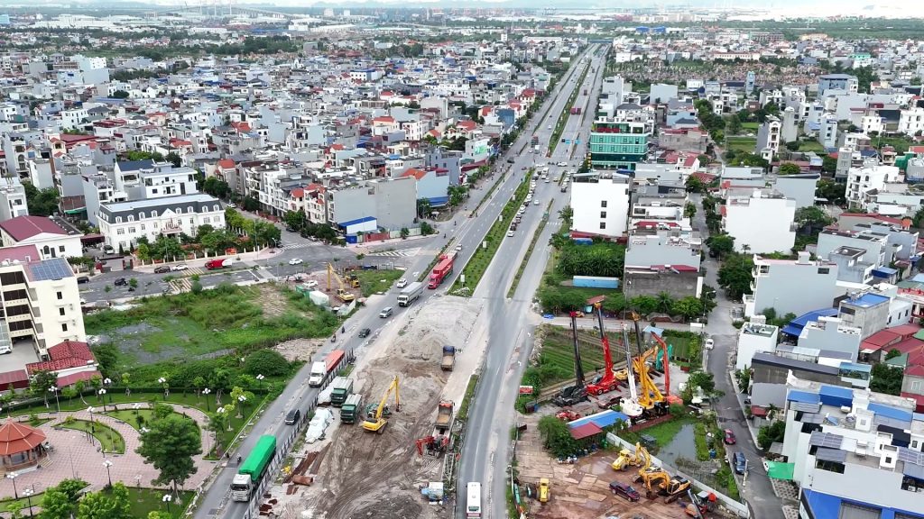 Trục đường lớn chạy thẳng qua khu dân cư đông đúc, đoạn giữa đang thi công với máy khoan cọc và xe tải, bên trái là một công viên nhỏ có chòi bát giác
