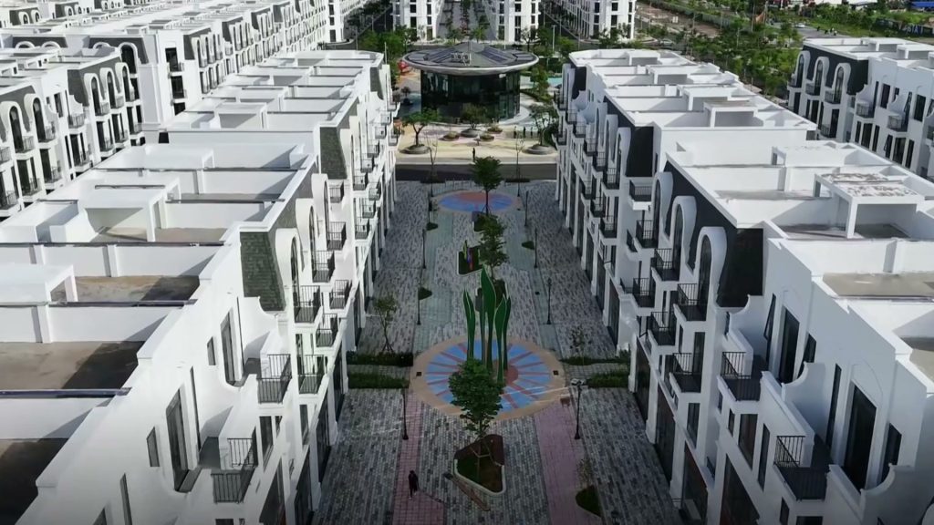 Hai dãy nhà phố mặt tiền trắng nhìn từ trên cao, giữa là quảng trường lát đá có bồn cây tròn và mái che hình vòm