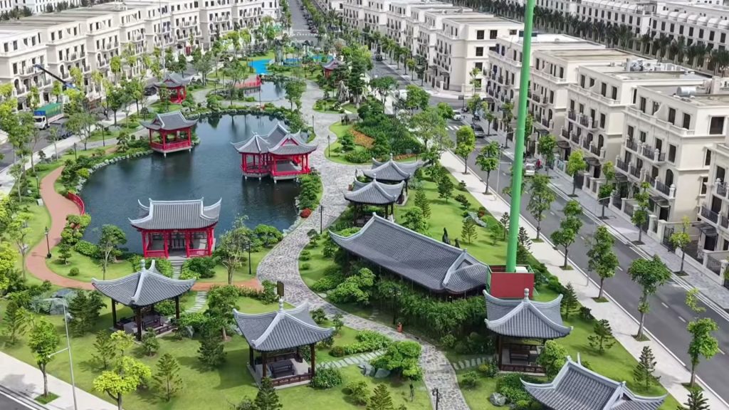 Vườn cảnh kiểu Á Đông trong khu đô thị trên đảo Vũ Yên nhìn từ trên cao, hồ nước có nhà thuỷ tạ mái đỏ, những mái ngói xám và lối đi lát đá giữa cây xanh, xung quanh là các dãy biệt thự trắng