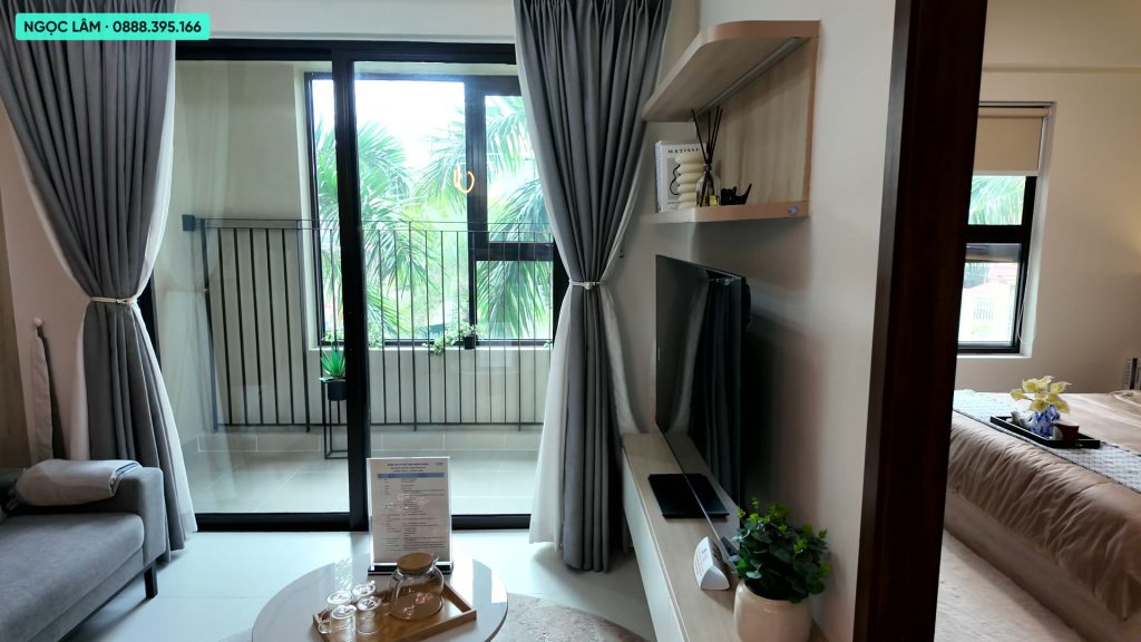 Phòng khách căn hộ mẫu 2 phòng ngủ AnZen Residences với sofa, kệ tivi gỗ sáng màu và cửa kính mở ra logia nhìn ra hàng cau