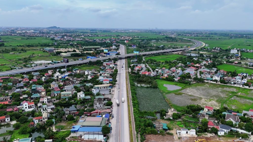Trục đường lớn chạy giữa khu dân cư và đồng ruộng, phía xa một tuyến cao tốc trên cao cắt ngang