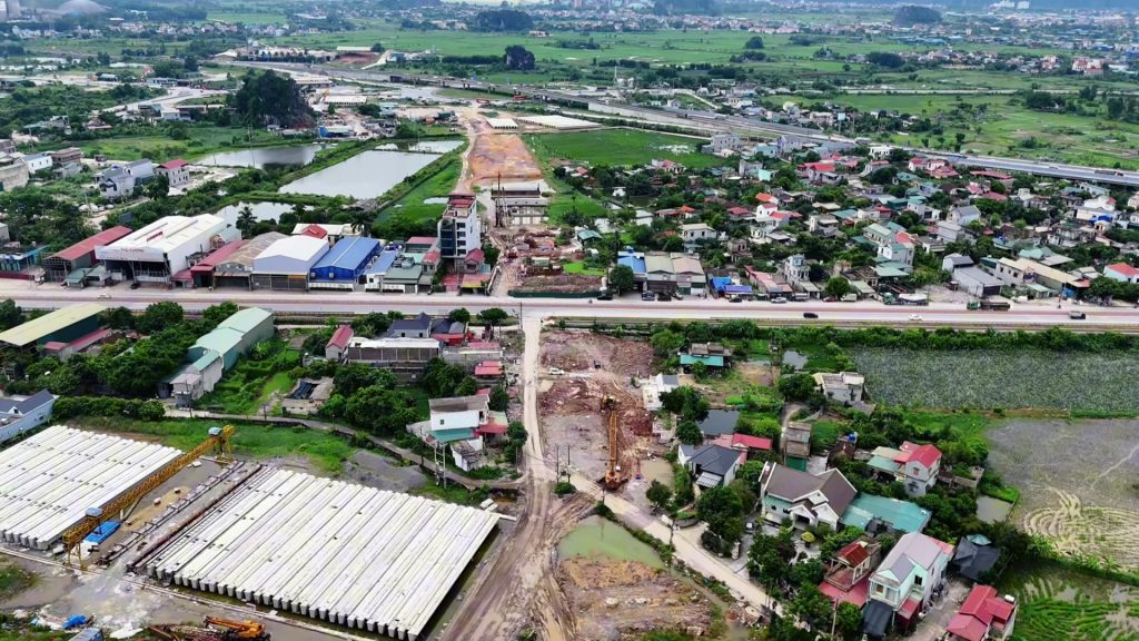 Công trường đường mới cắt ngang một quốc lộ, bên trái là bãi tập kết dầm cầu bê tông đúc sẵn và cần cẩu