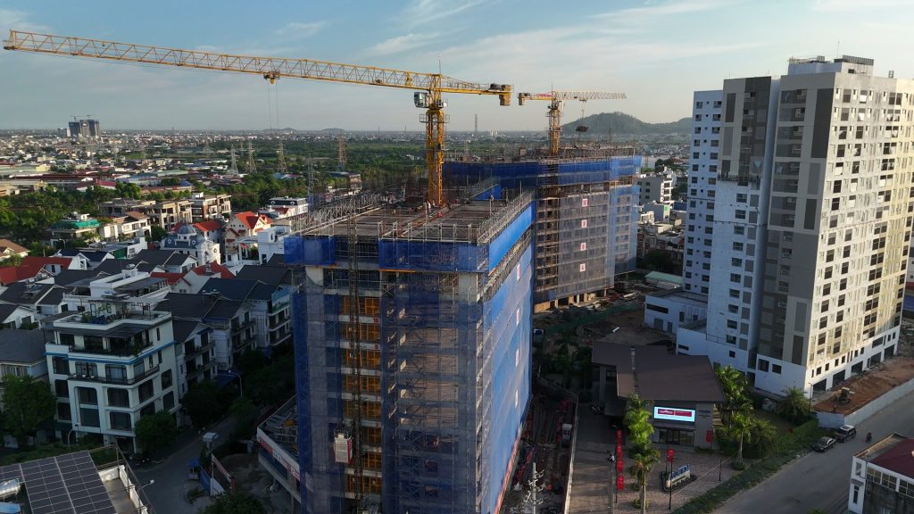 Hai tòa tháp AnZen Residences đang xây nhìn từ trên cao, giàn giáo phủ lưới xanh và hai cần cẩu tháp vàng, bên cạnh là tòa đã hoàn thiện màu trắng