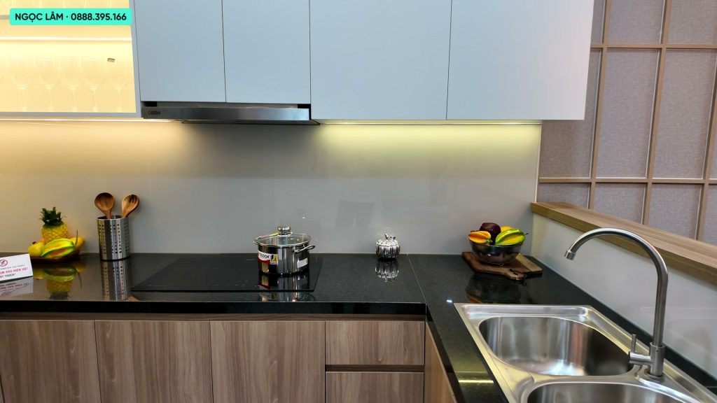Khu bếp căn hộ mẫu AnZen Residences với tủ bếp dưới gỗ vân sáng, mặt đá đen, bếp từ, máy hút mùi và bồn rửa hai ngăn
