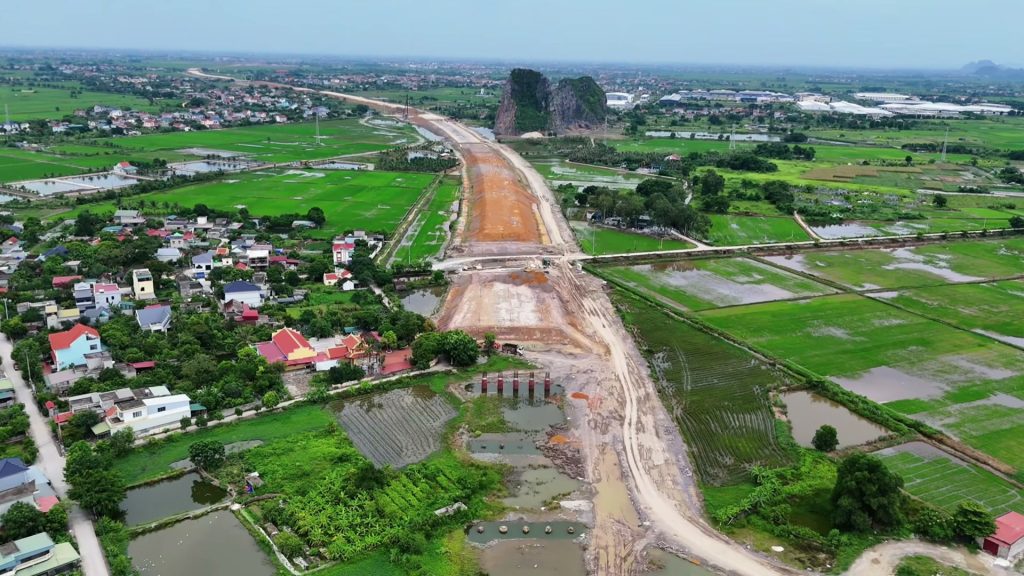 Nền đường cao tốc đang đắp chạy thẳng giữa những thửa ruộng, phía xa là một ngọn núi đá vôi và khu dân cư