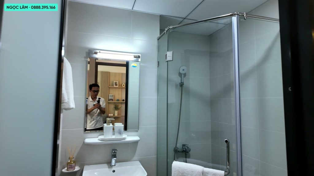 Nhà vệ sinh căn hộ mẫu AnZen Residences ốp gạch sáng màu, có buồng tắm kính, sen cây, chậu rửa treo tường và gương đèn